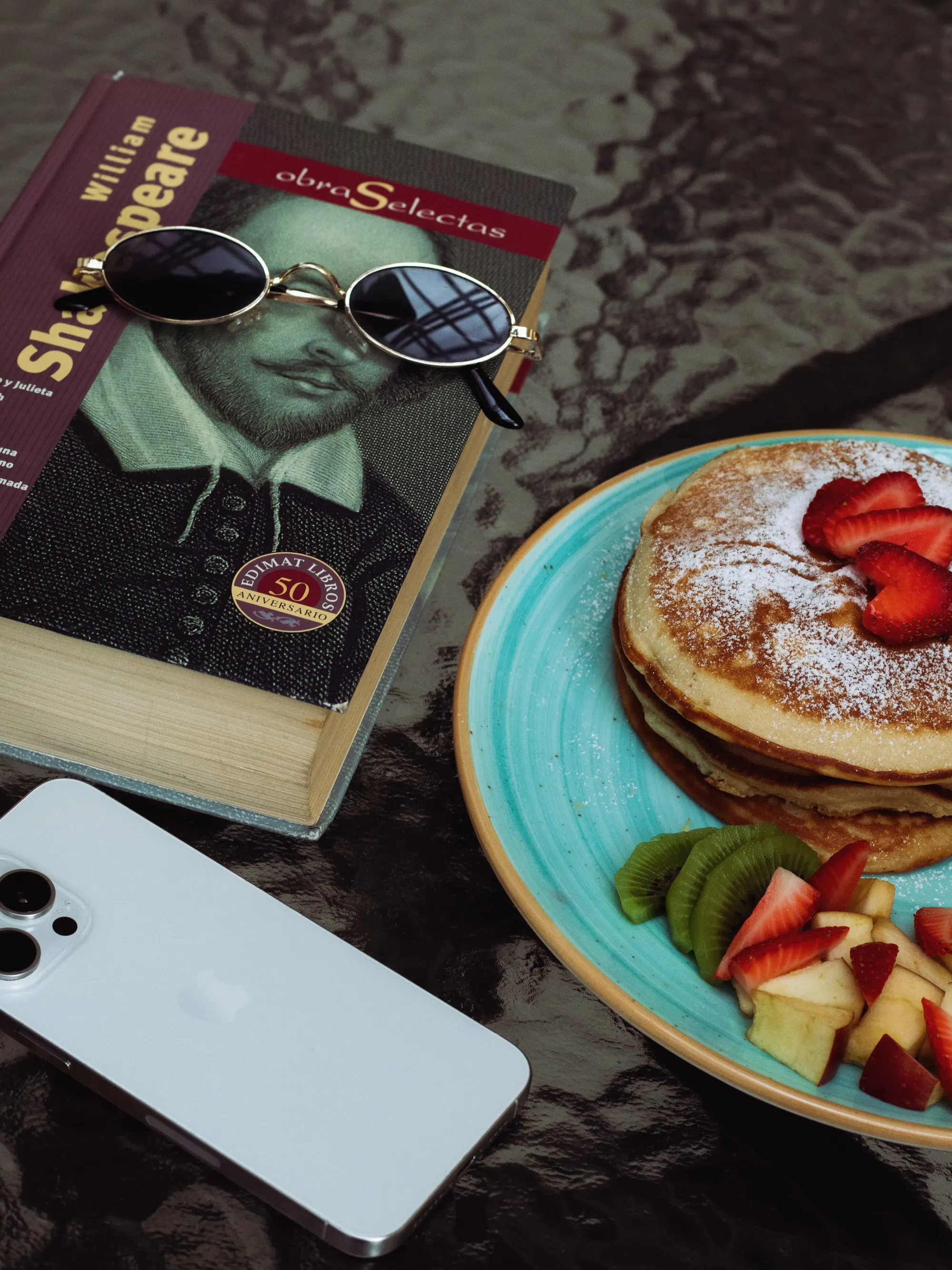 Pancakes con fresas y kiwi junto a un libro y unos lentes de sol