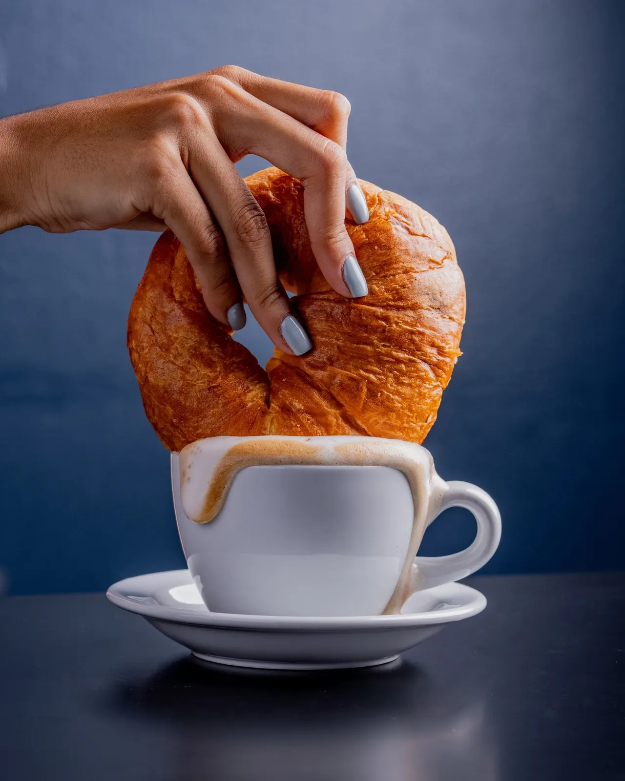 Una mano moja una concha en una taza de café frente a la pared azul